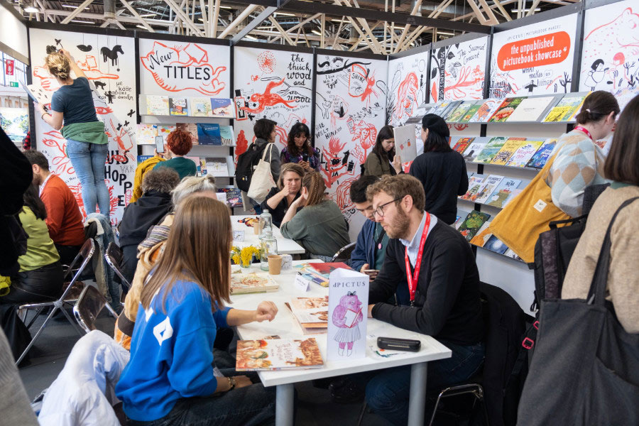 Hero image for Picture Book Makers featuring a photo of the organization's booth at a convention with people sitting at a table surrounded by a booth with books and illustrated walls.